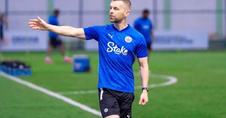 Image of Mumbai City FC Coach Petr Kratky, a man wearing a team tracksuit and cap, standing on a football field with a determined expression
