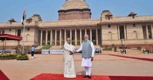 Indian Prime Minister Narendra Modi and Bangladesh Prime Minister Sheikh Hasina are seen smiling and shaking hands at a formal event. Both leaders are dressed in traditional attire; Modi is in a white kurta with a beige jacket, while Hasina is in a colorful saree."