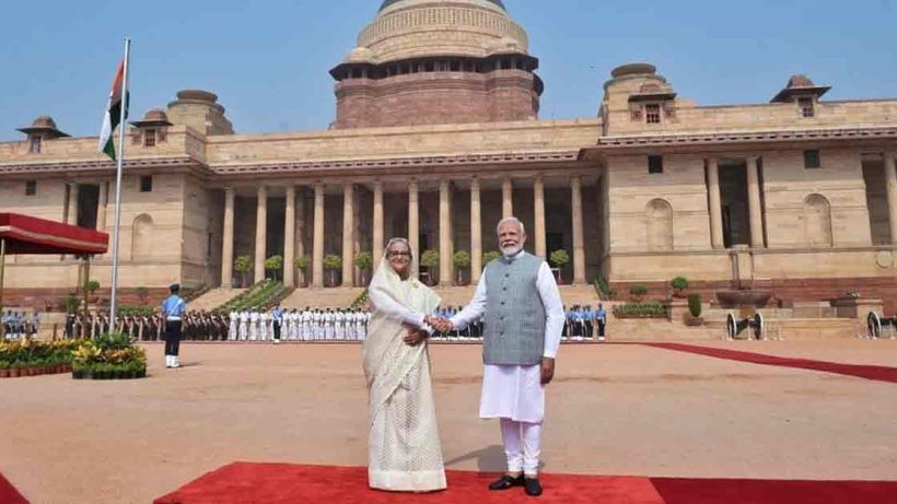 Indian Prime Minister Narendra Modi and Bangladesh Prime Minister Sheikh Hasina are seen smiling and shaking hands at a formal event. Both leaders are dressed in traditional attire; Modi is in a white kurta with a beige jacket, while Hasina is in a colorful saree."