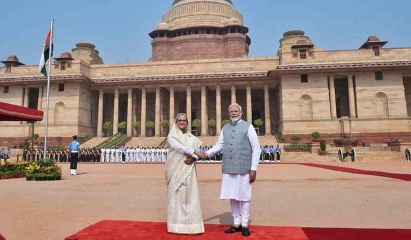 Indian Prime Minister Narendra Modi and Bangladesh Prime Minister Sheikh Hasina are seen smiling and shaking hands at a formal event. Both leaders are dressed in traditional attire; Modi is in a white kurta with a beige jacket, while Hasina is in a colorful saree."