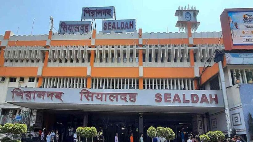 Sealdah Station in Kolkata, showing a bustling scene with passengers and trains. The station is large and busy, with numerous people walking and waiting on the platforms. The image captures the dynamic atmosphere of one of the city's major railway hubs.