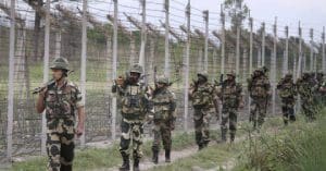 The image shows a group of Border Security Force (BSF) personnel standing in formation on a parade ground. They are dressed in khaki uniforms and distinctive headgear, with some holding rifles. The background features lush greenery and a clear blue sky.