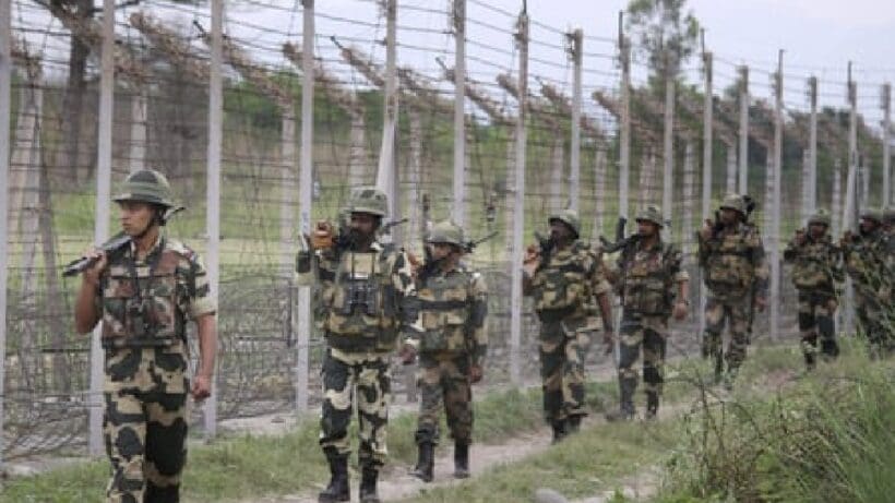 The image shows a group of Border Security Force (BSF) personnel standing in formation on a parade ground. They are dressed in khaki uniforms and distinctive headgear, with some holding rifles. The background features lush greenery and a clear blue sky.