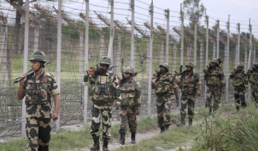 The image shows a group of Border Security Force (BSF) personnel standing in formation on a parade ground. They are dressed in khaki uniforms and distinctive headgear, with some holding rifles. The background features lush greenery and a clear blue sky.