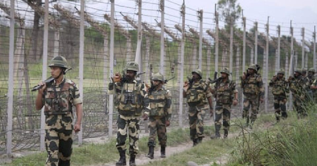 The image shows a group of Border Security Force (BSF) personnel standing in formation on a parade ground. They are dressed in khaki uniforms and distinctive headgear, with some holding rifles. The background features lush greenery and a clear blue sky.