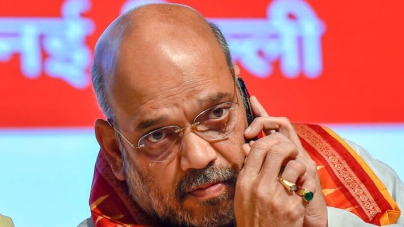 Amit Shah, Union Home Minister of India, wearing a traditional white kurta and a saffron scarf, is smiling and posing for the camera in a well-lit setting.