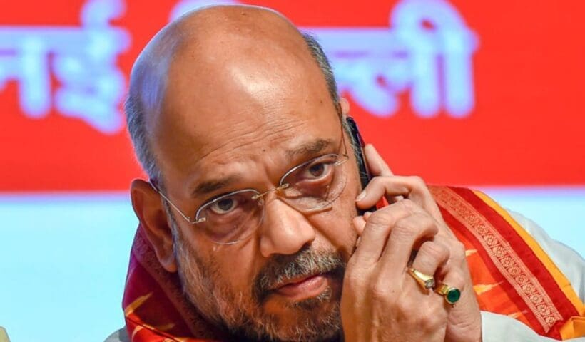 Amit Shah, Union Home Minister of India, wearing a traditional white kurta and a saffron scarf, is smiling and posing for the camera in a well-lit setting.