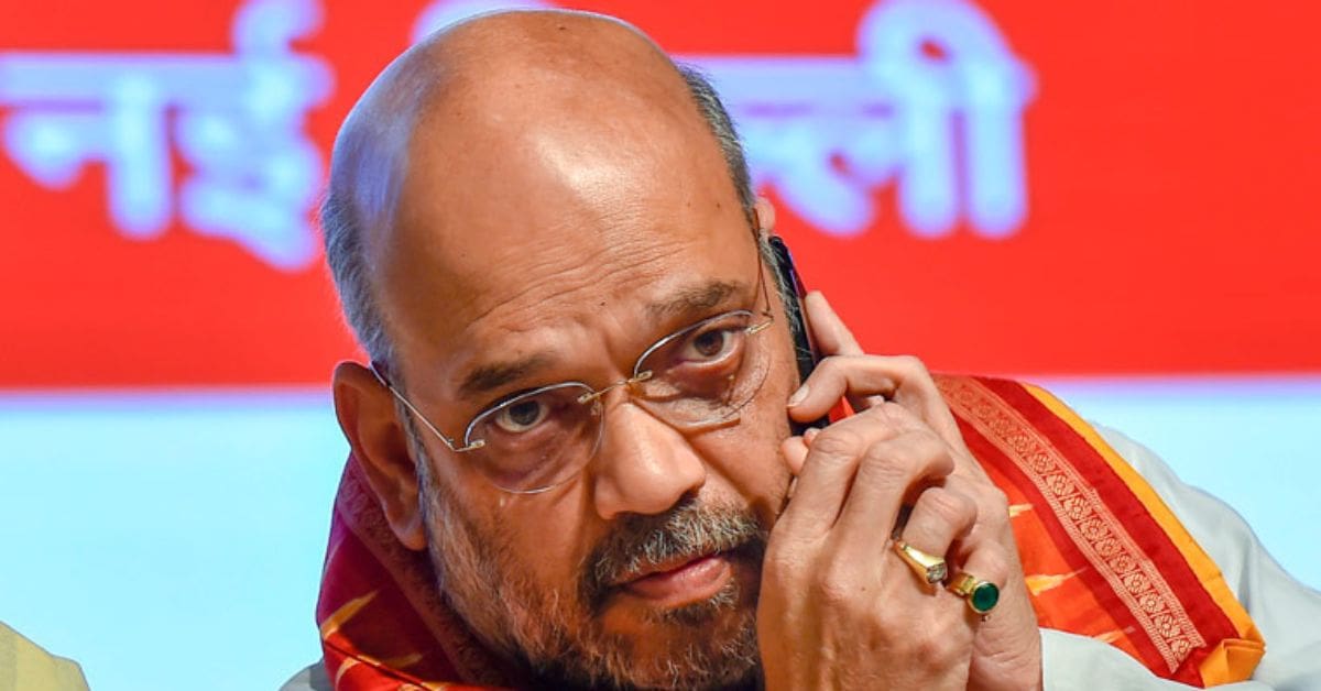 Amit Shah, Union Home Minister of India, wearing a traditional white kurta and a saffron scarf, is smiling and posing for the camera in a well-lit setting.