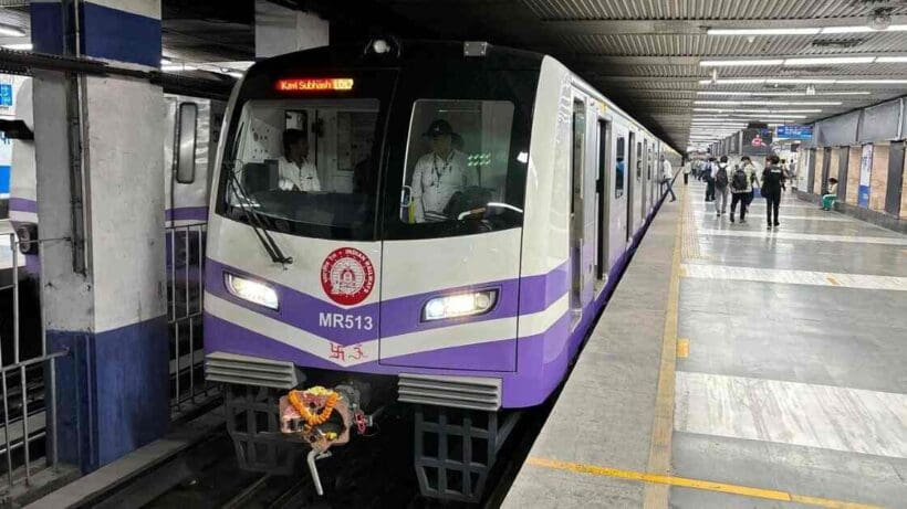 Image of a Kolkata Metro train on a track, showcasing a modern, sleek design with a prominent blue and white color scheme. The train is positioned in an elevated metro station with visible city buildings in the background.