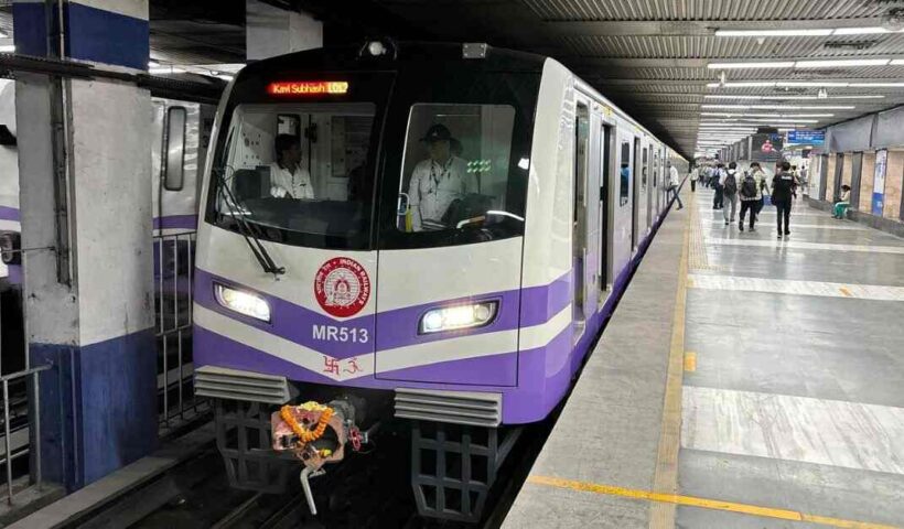 Image of a Kolkata Metro train on a track, showcasing a modern, sleek design with a prominent blue and white color scheme. The train is positioned in an elevated metro station with visible city buildings in the background.