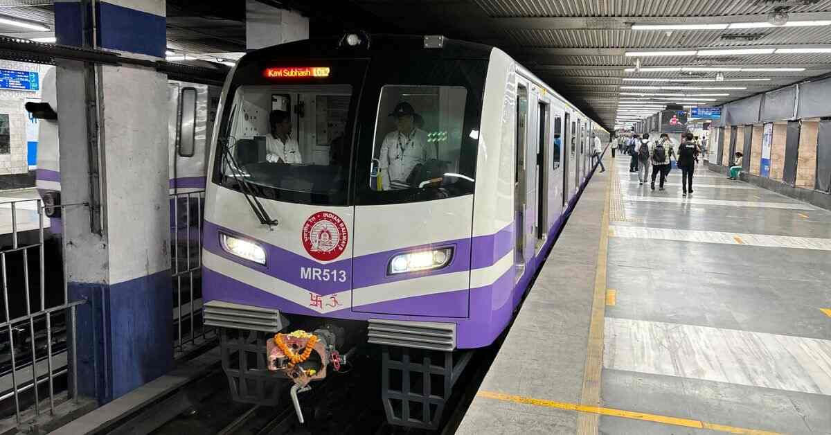 Image of a Kolkata Metro train on a track, showcasing a modern, sleek design with a prominent blue and white color scheme. The train is positioned in an elevated metro station with visible city buildings in the background.