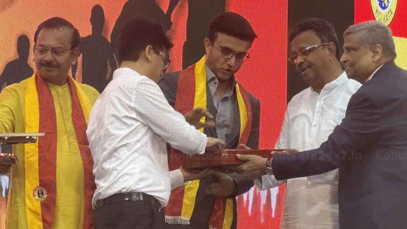 Sourav Ganguly, the renowned Indian cricketer, shares his thoughts on the iconic East Bengal football jersey. The image features Ganguly holding the East Bengal jersey, smiling and seated in a well-lit room with sports memorabilia in the background.