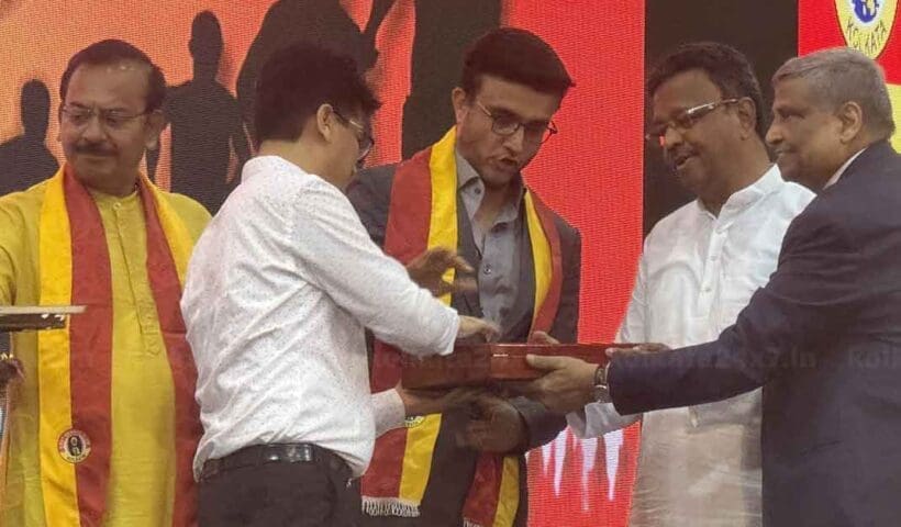 Sourav Ganguly, the renowned Indian cricketer, shares his thoughts on the iconic East Bengal football jersey. The image features Ganguly holding the East Bengal jersey, smiling and seated in a well-lit room with sports memorabilia in the background.