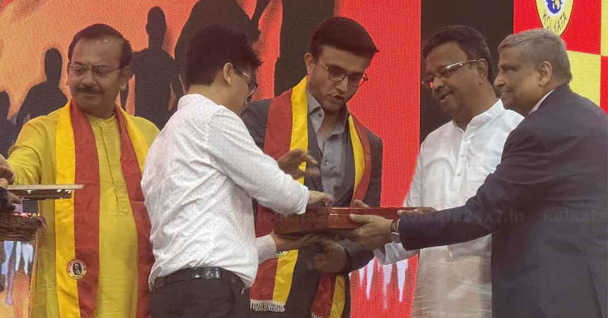 Sourav Ganguly, the renowned Indian cricketer, shares his thoughts on the iconic East Bengal football jersey. The image features Ganguly holding the East Bengal jersey, smiling and seated in a well-lit room with sports memorabilia in the background.