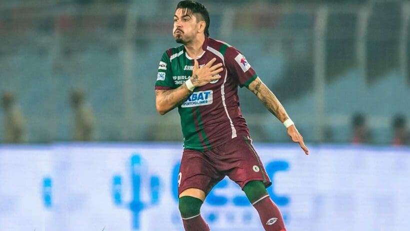 Dimitri Petratos, the Australian footballer, is pictured in his Mohun Bagan SG jersey. He is standing confidently on a football field, with stadium lights in the background, highlighting his presence on the team.