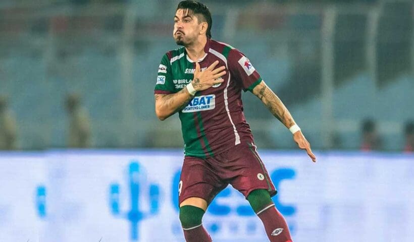 Dimitri Petratos, the Australian footballer, is pictured in his Mohun Bagan SG jersey. He is standing confidently on a football field, with stadium lights in the background, highlighting his presence on the team.