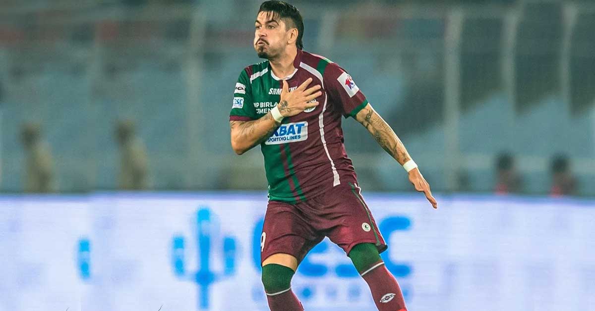 Dimitri Petratos, the Australian footballer, is pictured in his Mohun Bagan SG jersey. He is standing confidently on a football field, with stadium lights in the background, highlighting his presence on the team.